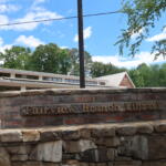 Stone brick sign wall with carved text reading 'Fairview Branch Library' in front of a low building and green trees under a blue sky.