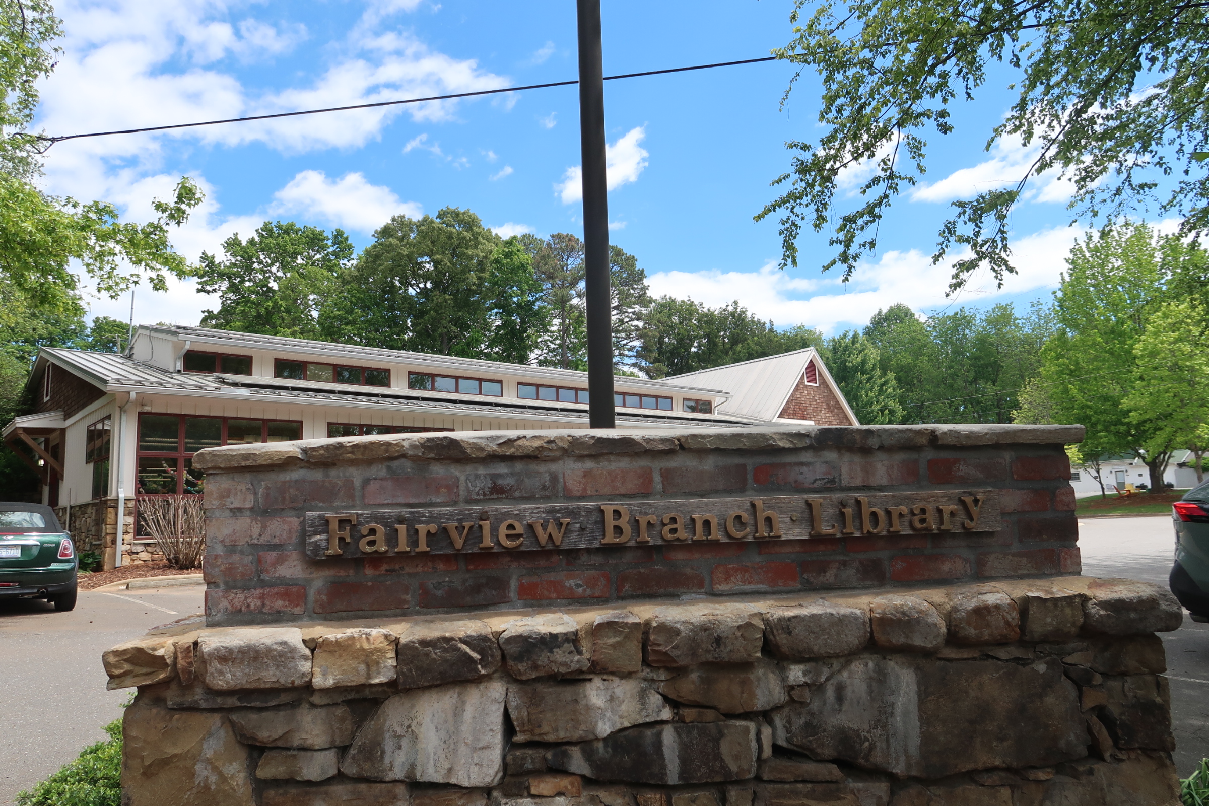 Stone brick sign wall with carved text reading 'Fairview Branch Library' in front of a low building and green trees under a blue sky.