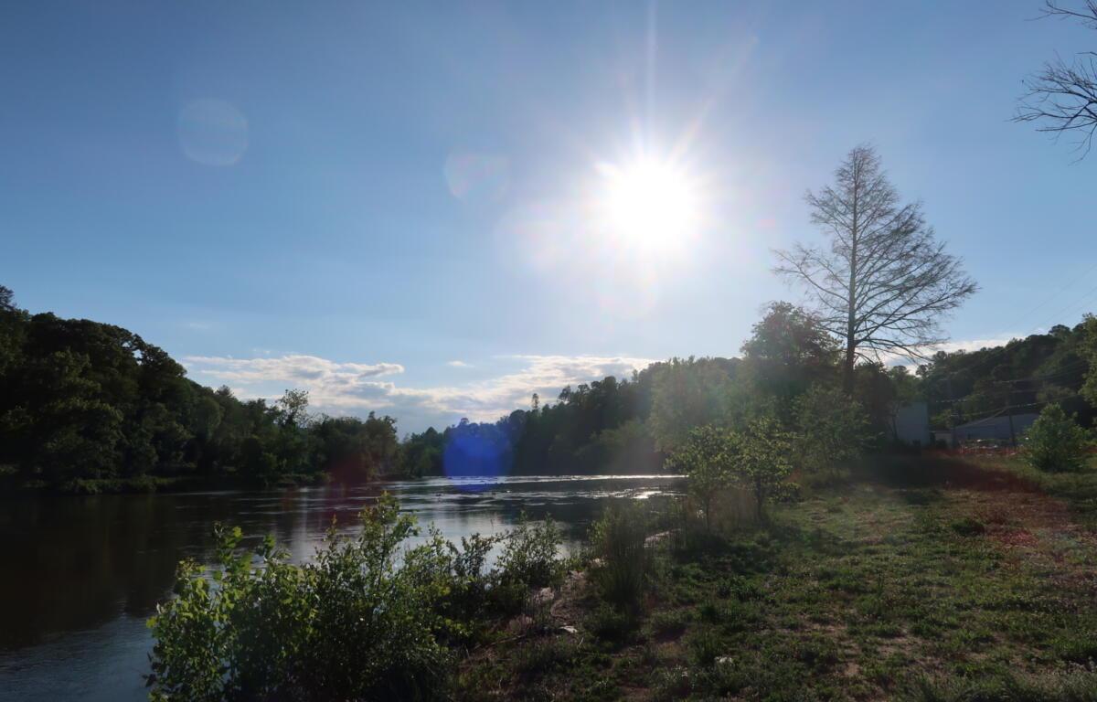 Bright sunlit river scene with a clear blue sky, trees along the banks, and a calm water surface below.