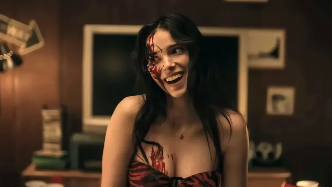 Young woman in a red-and-black dress, blood on her face and chest, smiling in a dimly lit room.