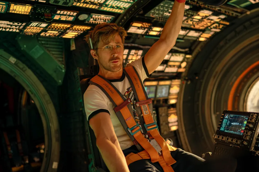 Pilot in a high-tech aircraft cockpit, wearing an orange harness and reaching up toward the overhead controls.