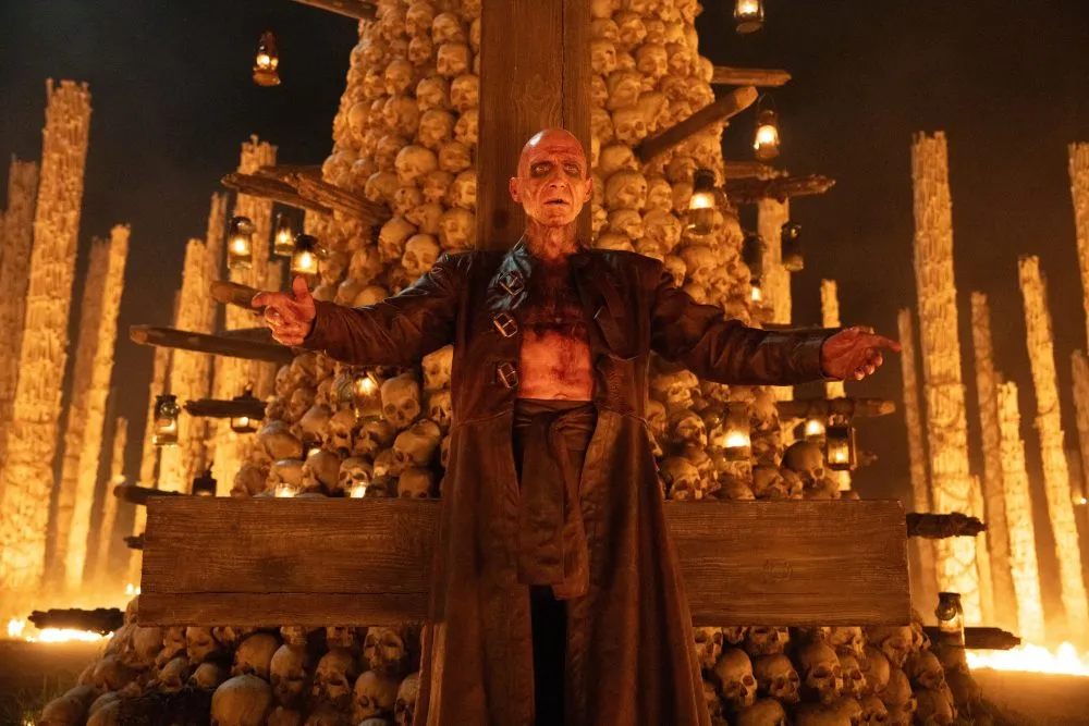 Pale, bald man in a long brown coat stands with arms outstretched before a towering stack of skulls lit by torches.