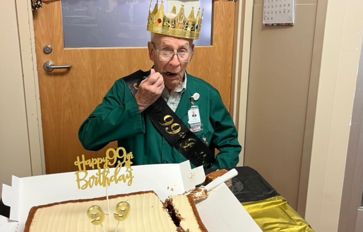 Elderly man wearing a golden crown and birthday sash, about to eat cake at a 99th birthday celebration indoors.
