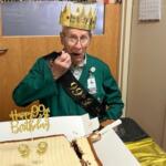 Elderly man wearing a golden crown and birthday sash, about to eat cake at a 99th birthday celebration indoors.