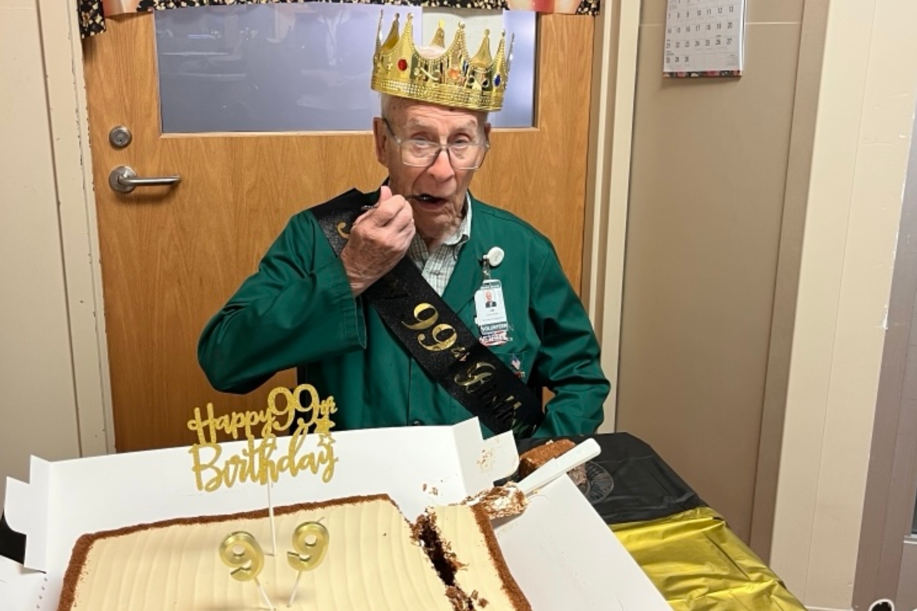 Elderly man wearing a golden crown and birthday sash, about to eat cake at a 99th birthday celebration indoors.