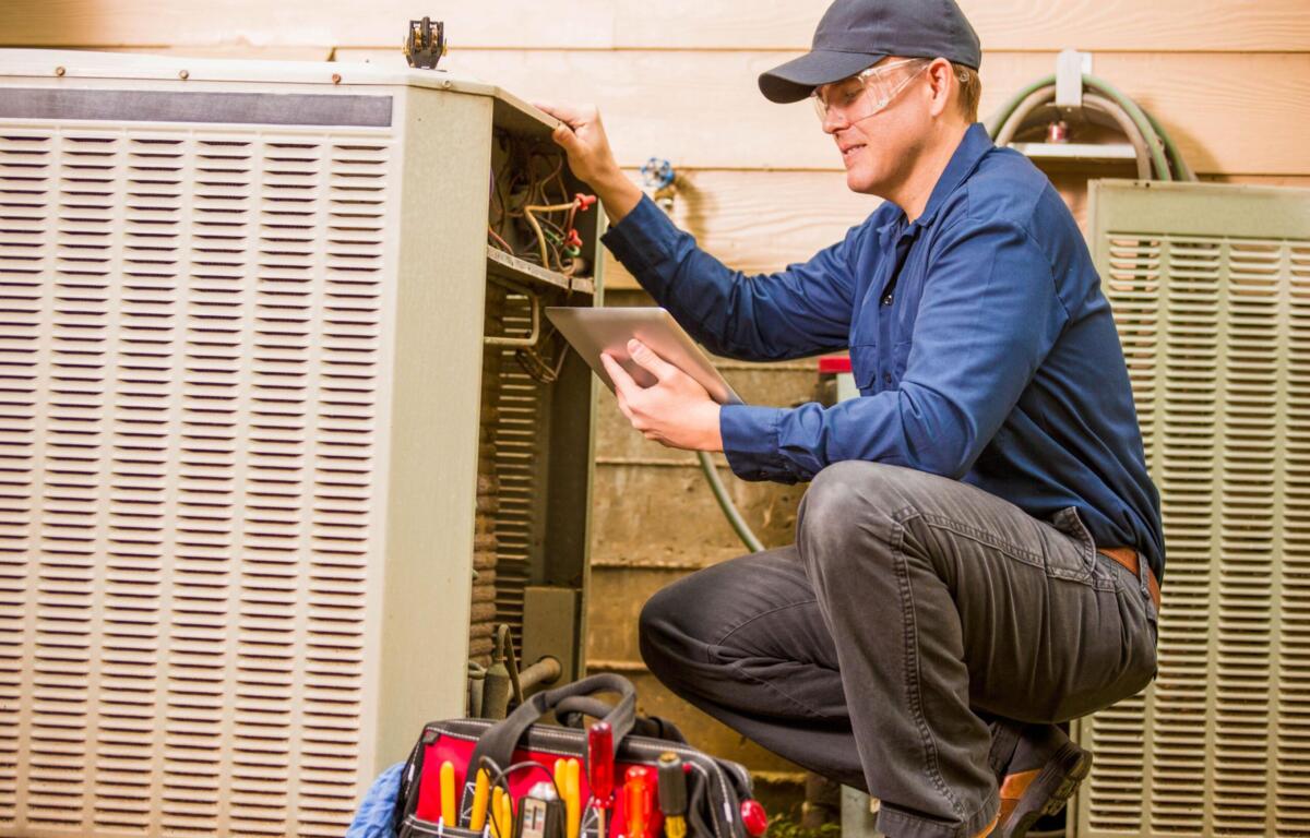 HVAC technician wearing a cap and safety glasses kneels by an outdoor air conditioner, inspecting wiring with a tablet.