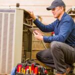 HVAC technician wearing a cap and safety glasses kneels by an outdoor air conditioner, inspecting wiring with a tablet.