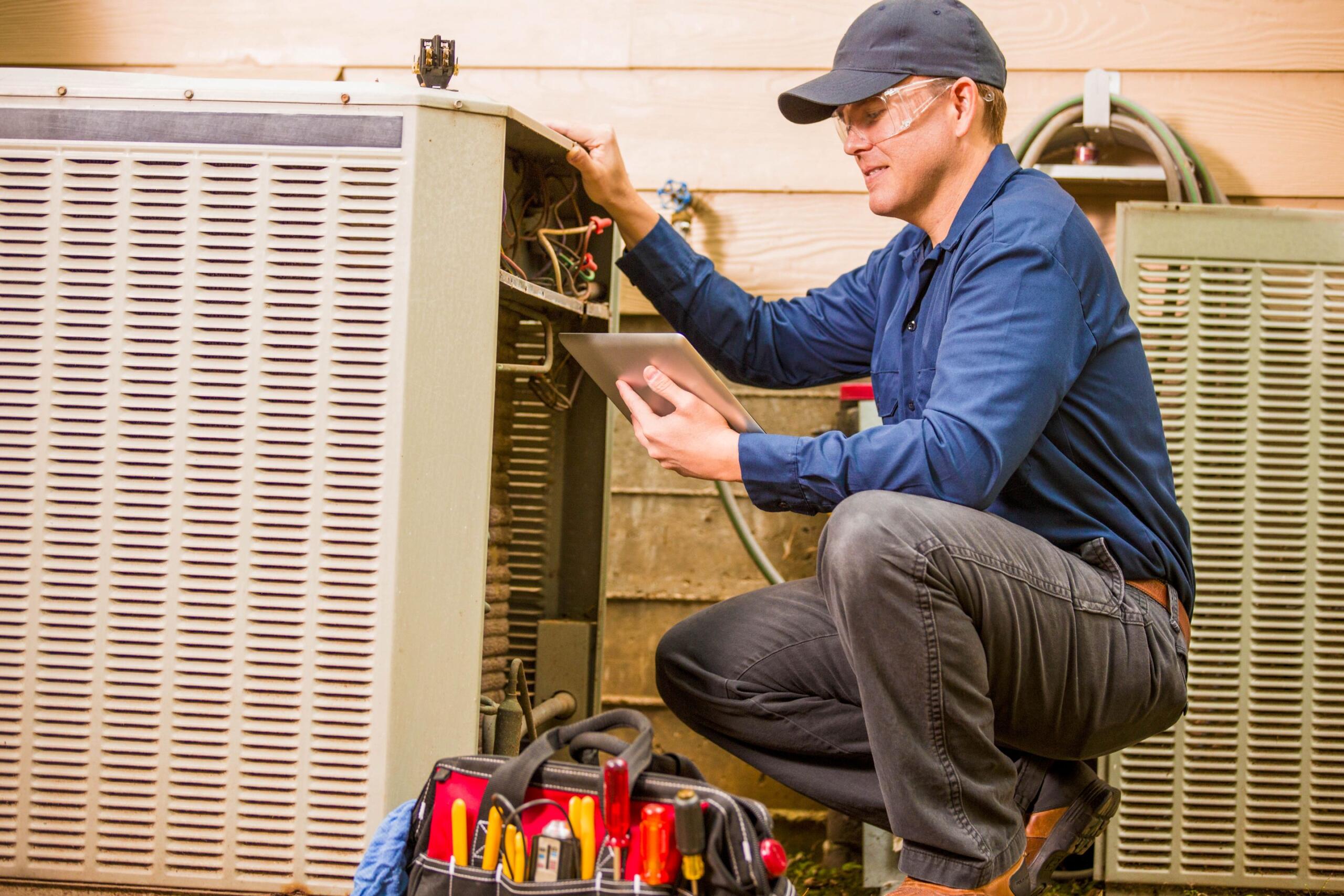 HVAC technician wearing a cap and safety glasses kneels by an outdoor air conditioner, inspecting wiring with a tablet.