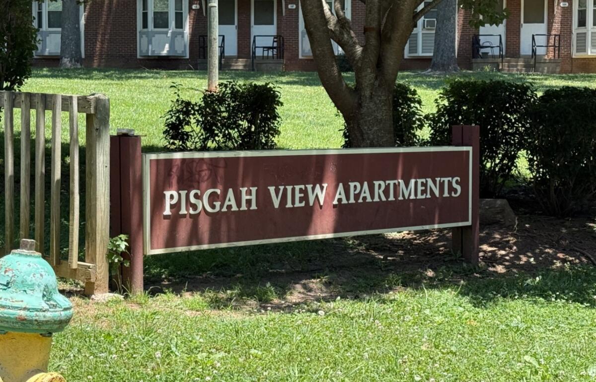 Sign for Pisgah View Apartments on a maroon board in front of a brick apartment building and greenery.