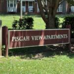 Sign for Pisgah View Apartments on a maroon board in front of a brick apartment building and greenery.