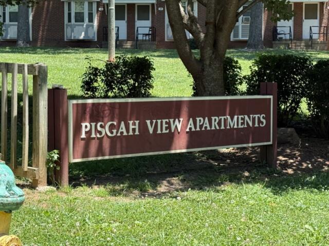 Sign for Pisgah View Apartments on a maroon board in front of a brick apartment building and greenery.
