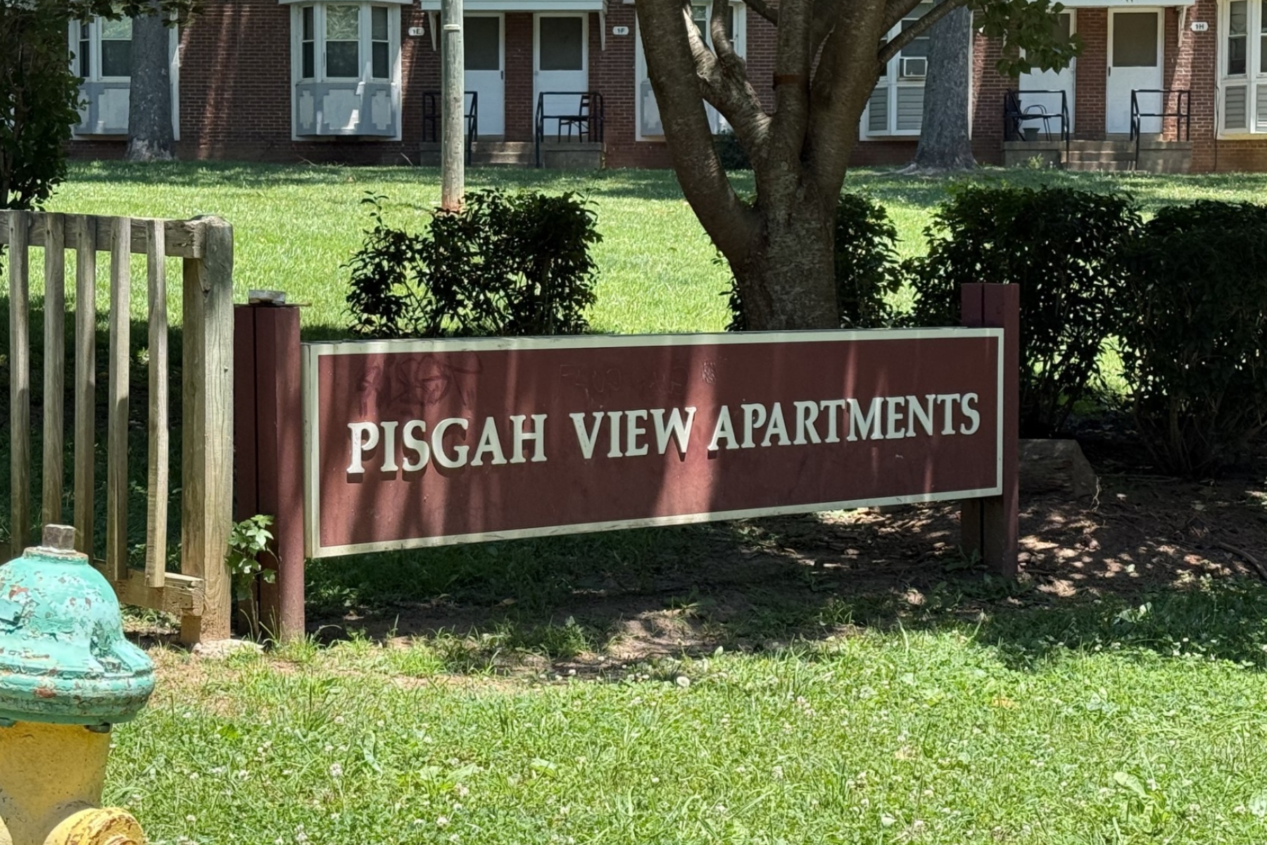 Sign for Pisgah View Apartments on a maroon board in front of a brick apartment building and greenery.