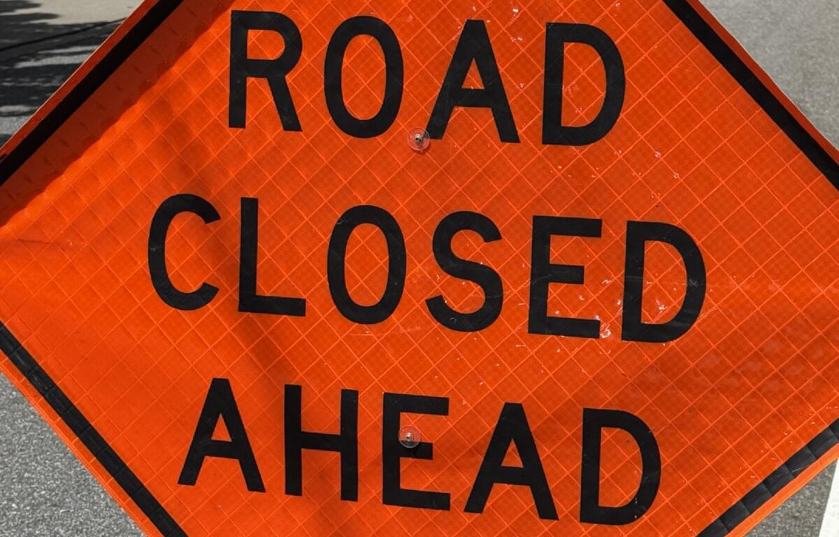 Orange diamond traffic sign with bold black text: 'ROAD CLOSED AHEAD' on pavement, close-up poster-style warning.