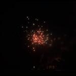 Night scene of red and white fireworks exploding above a dark neighborhood, with silhouettes of houses and spectators at ground level.