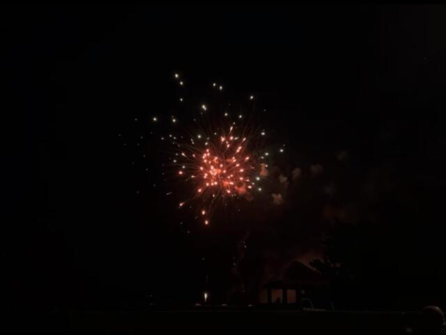 Night scene of red and white fireworks exploding above a dark neighborhood, with silhouettes of houses and spectators at ground level.