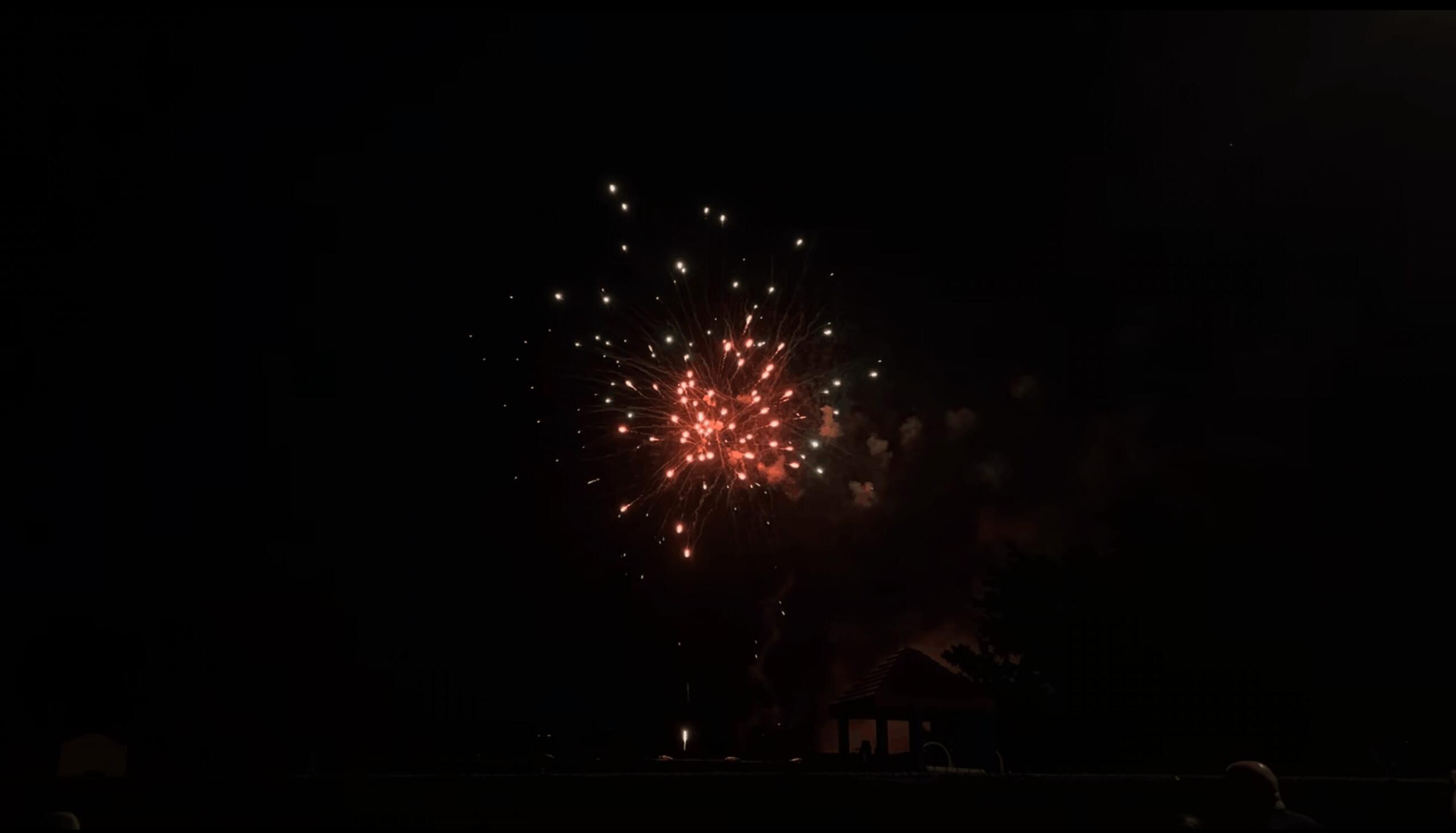 Night scene of red and white fireworks exploding above a dark neighborhood, with silhouettes of houses and spectators at ground level.