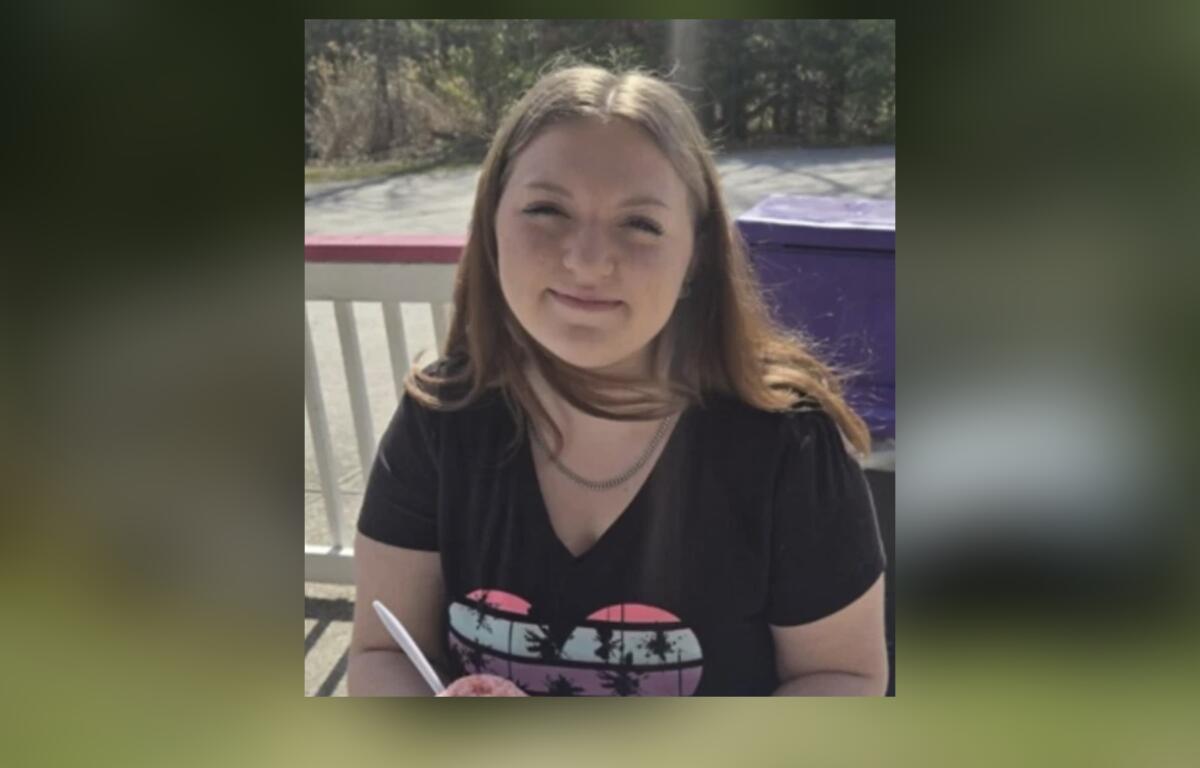 Young woman with long light brown hair wearing a black T‑shirt, outdoors on a porch, holding a pink ice cream cone.