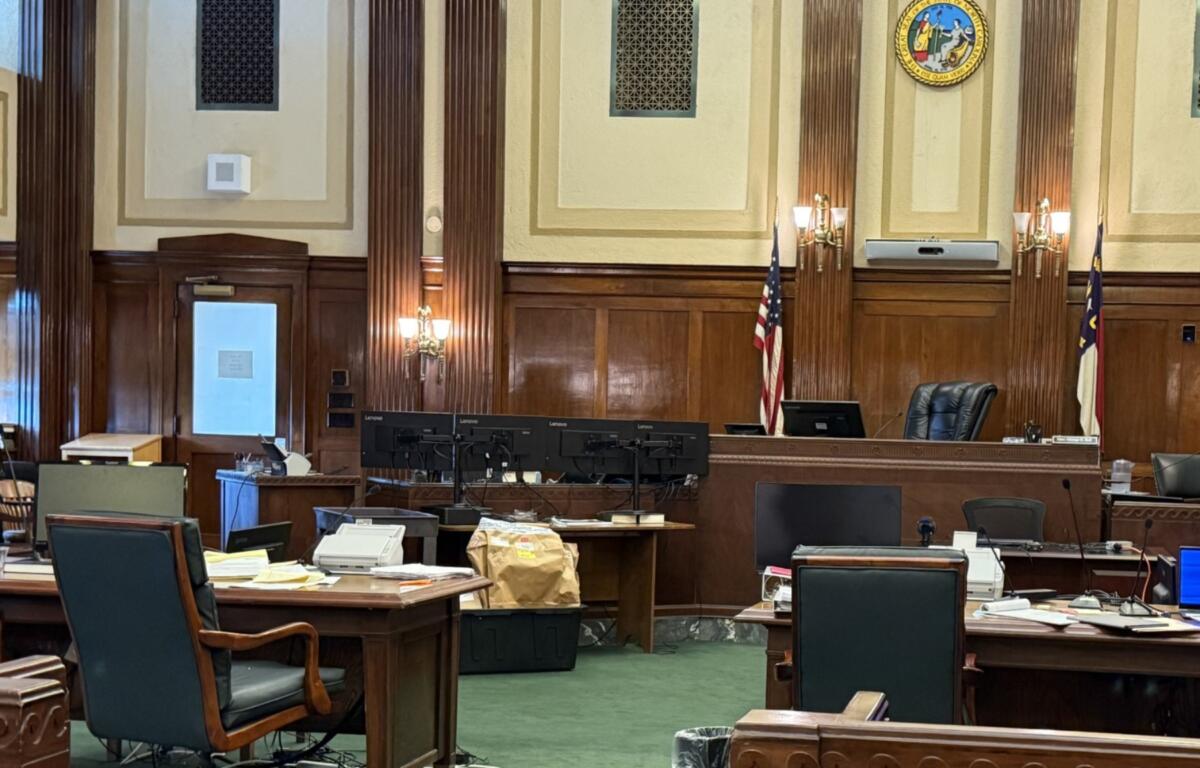 Courtroom or legislative chamber with wooden desks, monitors, and chairs arranged around a central bench; U.S. flag and state flag visible.