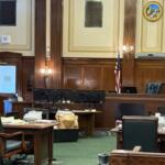 Courtroom or legislative chamber with wooden desks, monitors, and chairs arranged around a central bench; U.S. flag and state flag visible.