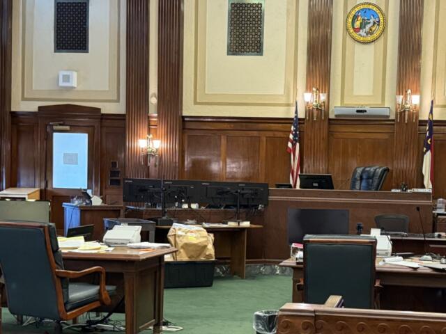 Courtroom or legislative chamber with wooden desks, monitors, and chairs arranged around a central bench; U.S. flag and state flag visible.