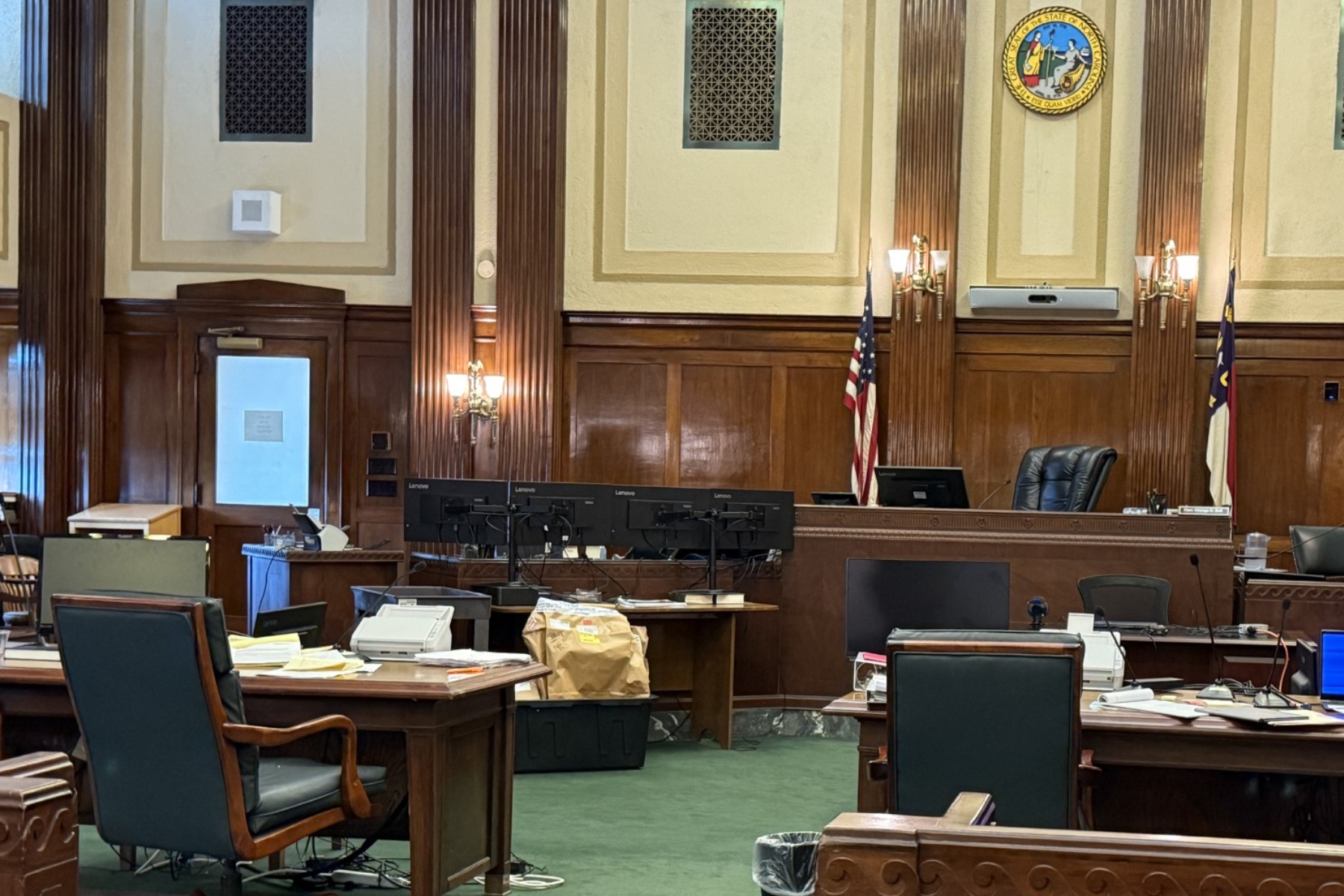 Courtroom or legislative chamber with wooden desks, monitors, and chairs arranged around a central bench; U.S. flag and state flag visible.