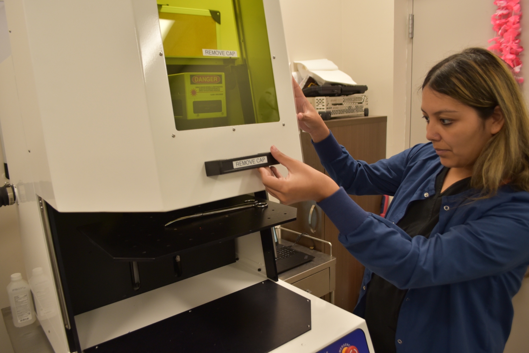 Person in a blue lab coat operates a large white lab instrument, grasping a labeled 'REMOVE CAP' handle.