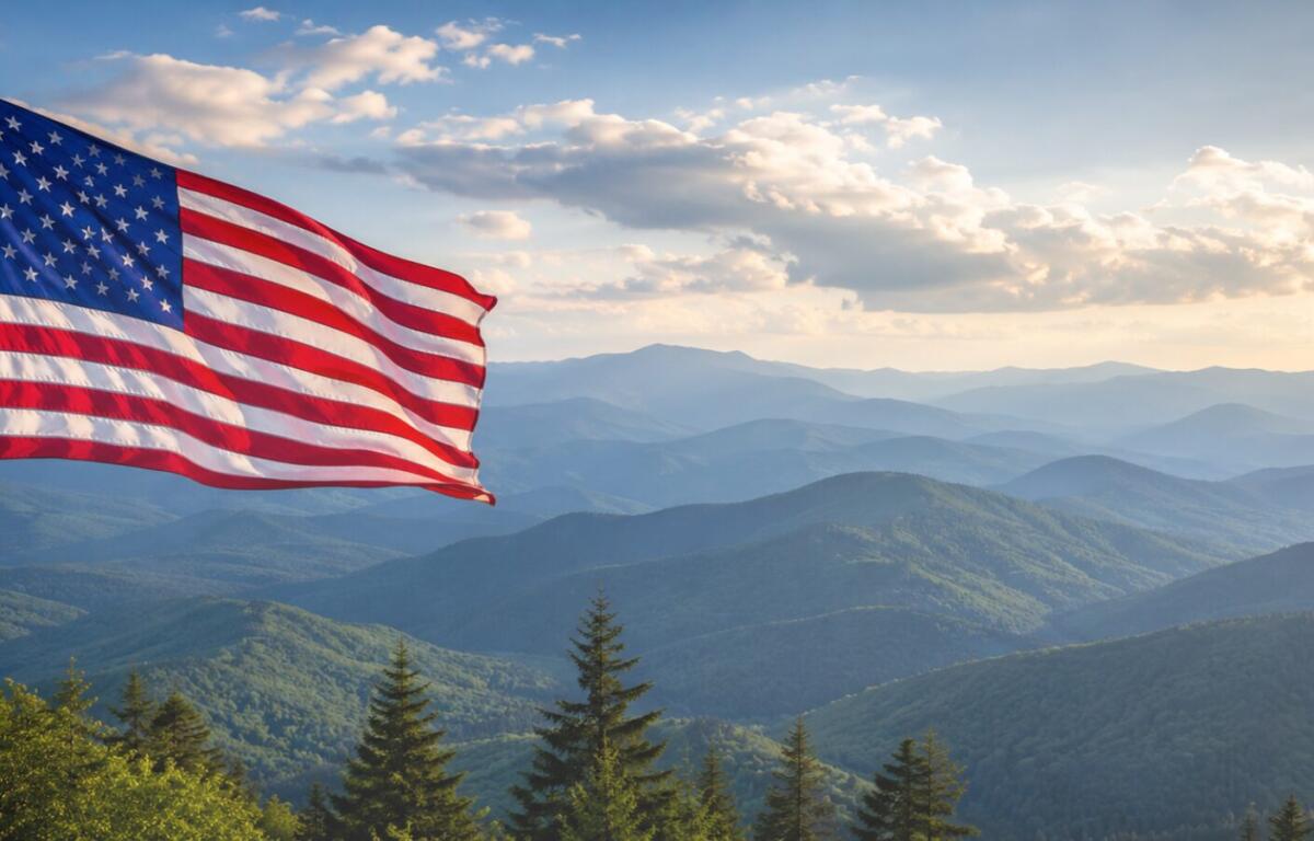 American flag waving on a flagpole with blue mountain ranges and evergreen trees in the foreground under a partly cloudy sky.