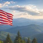 American flag waving on a flagpole with blue mountain ranges and evergreen trees in the foreground under a partly cloudy sky.