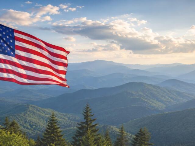 American flag waving on a flagpole with blue mountain ranges and evergreen trees in the foreground under a partly cloudy sky.