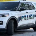 White police SUV parked near a building with yellow police tape across the scene, 'POLICE' lettering visible on the side.