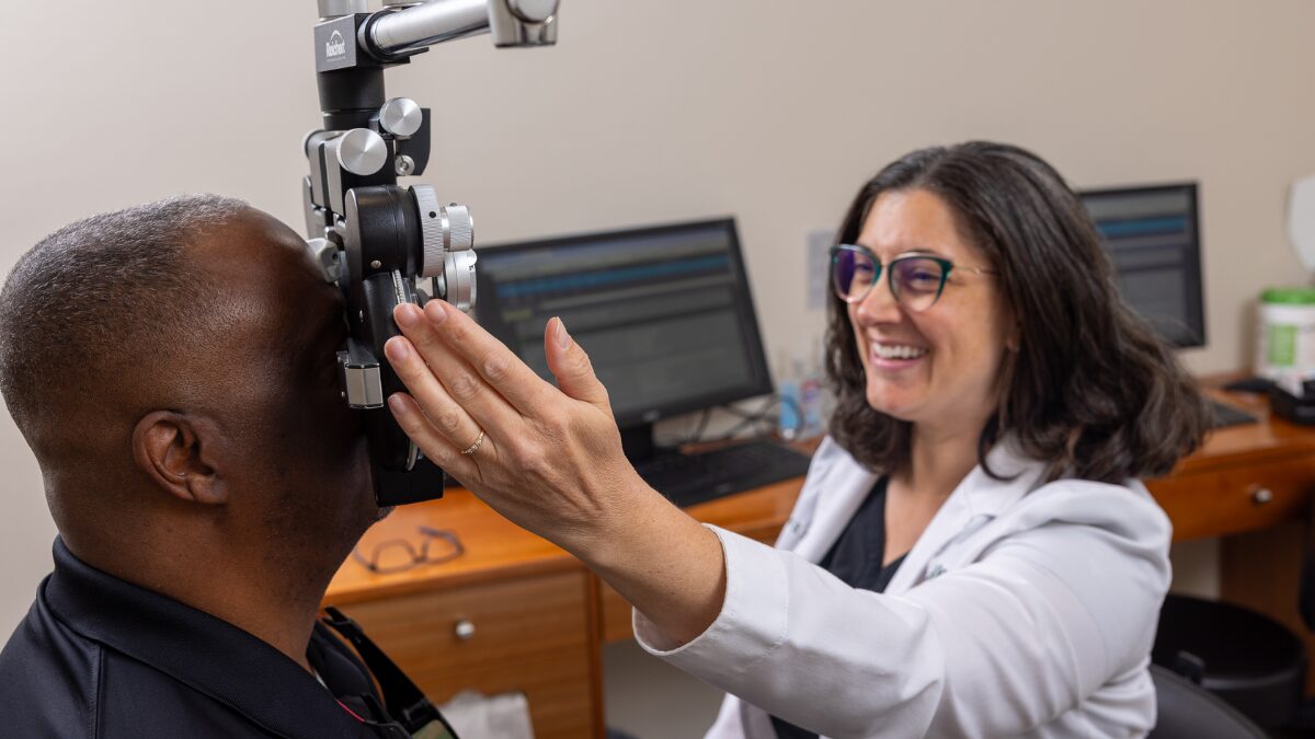 Optometrist adjusts a phoropter to test a patient's vision, both smiling in a clinic room.