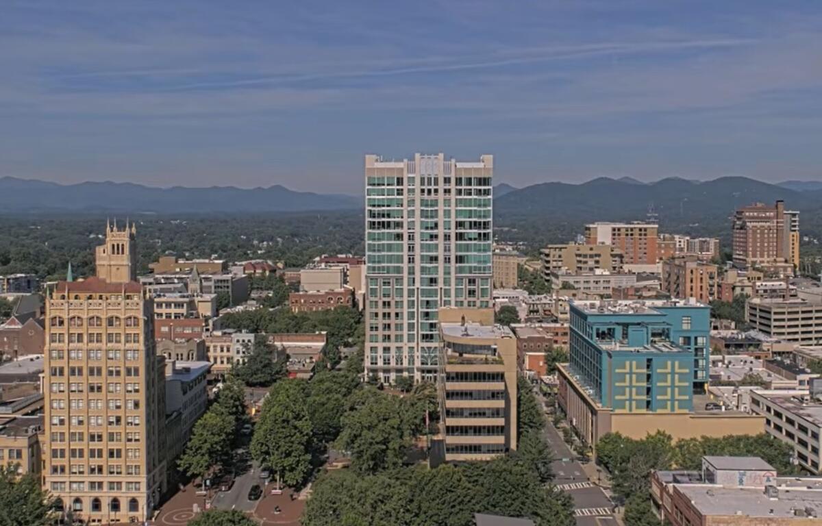 A downtown cityscape with a tall glass-fronted high-rise in the center, surrounded by midrise brick buildings and trees, mountains in the distance under a blue sky.
