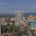 A downtown cityscape with a tall glass-fronted high-rise in the center, surrounded by midrise brick buildings and trees, mountains in the distance under a blue sky.