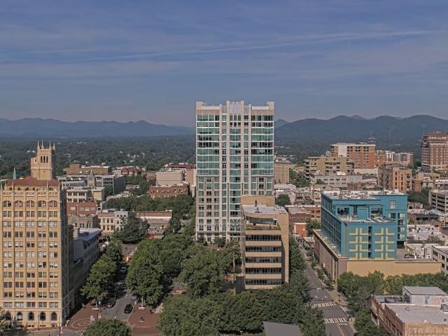 A downtown cityscape with a tall glass-fronted high-rise in the center, surrounded by midrise brick buildings and trees, mountains in the distance under a blue sky.