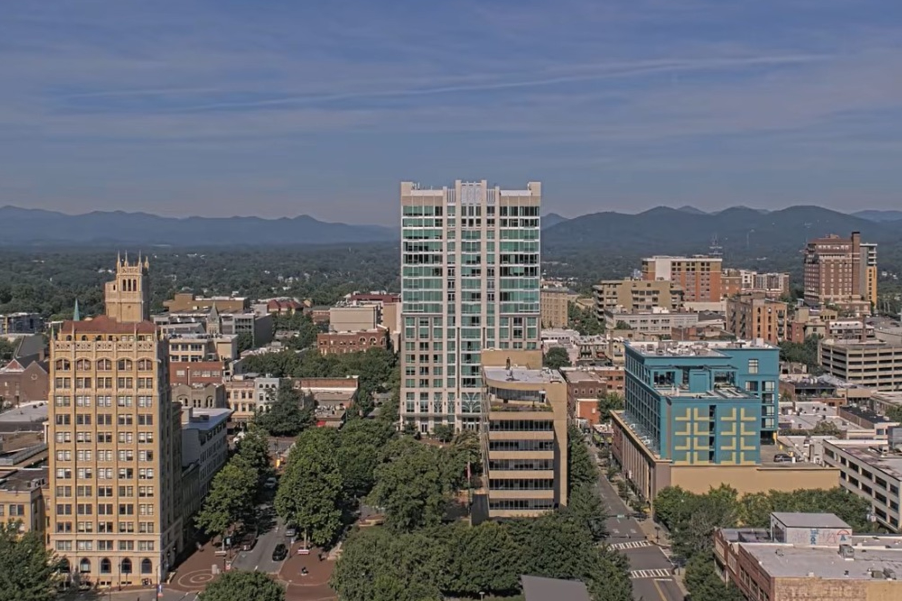 A downtown cityscape with a tall glass-fronted high-rise in the center, surrounded by midrise brick buildings and trees, mountains in the distance under a blue sky.