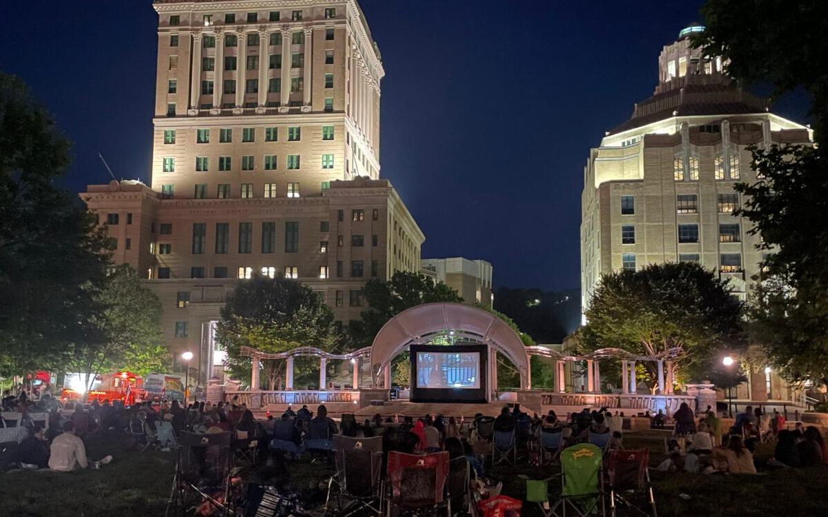 Nighttime outdoor movie screening in a city park with a large screen, audience on blankets, and tall illuminated buildings in the background.