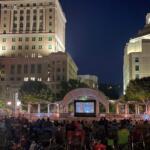 Nighttime outdoor movie screening in a city park with a large screen, audience on blankets, and tall illuminated buildings in the background.