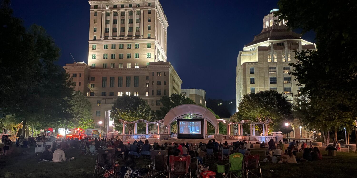 Nighttime outdoor movie screening in a city park with a large screen, audience on blankets, and tall illuminated buildings in the background.