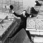 Man in a suit hanging from a rooftop ledge, tipping his hat toward the camera as he balances on the edge (black-and-white historic photo).