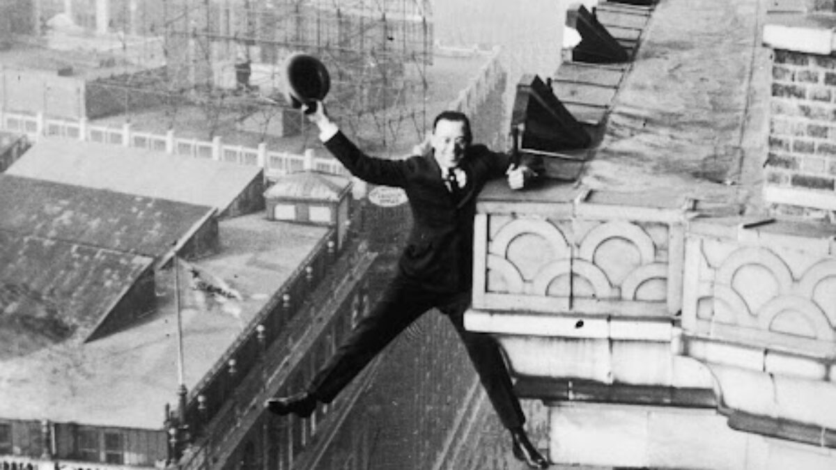 Man in a suit hanging from a rooftop ledge, tipping his hat toward the camera as he balances on the edge (black-and-white historic photo).