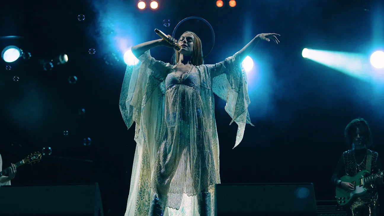 Female singer on stage in a shimmering cape dress, singing into a microphone with blue spotlights behind her.
