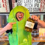 Man in a bright green pickle costume points to a jar labeled “PICKLES MAKE GREAT BEER” at a bar with beer taps in the background, smiling widely.