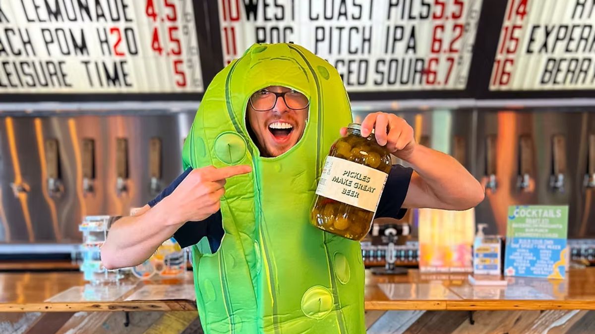 Man in a bright green pickle costume points to a jar labeled “PICKLES MAKE GREAT BEER” at a bar with beer taps in the background, smiling widely.