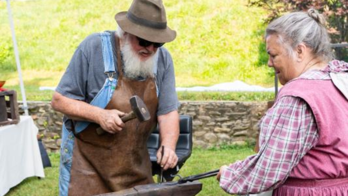 Two older adults outside, man with a beard and hat hammering metal while a woman in a pink vest watches nearby.