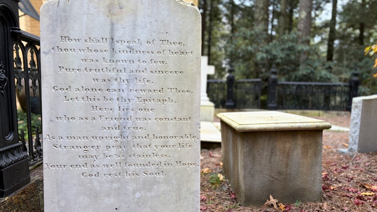 Stone tombstone with an engraved poem in a cemetery; the visible lines begin 'How shall I speak of Thee...' with trees in the background.