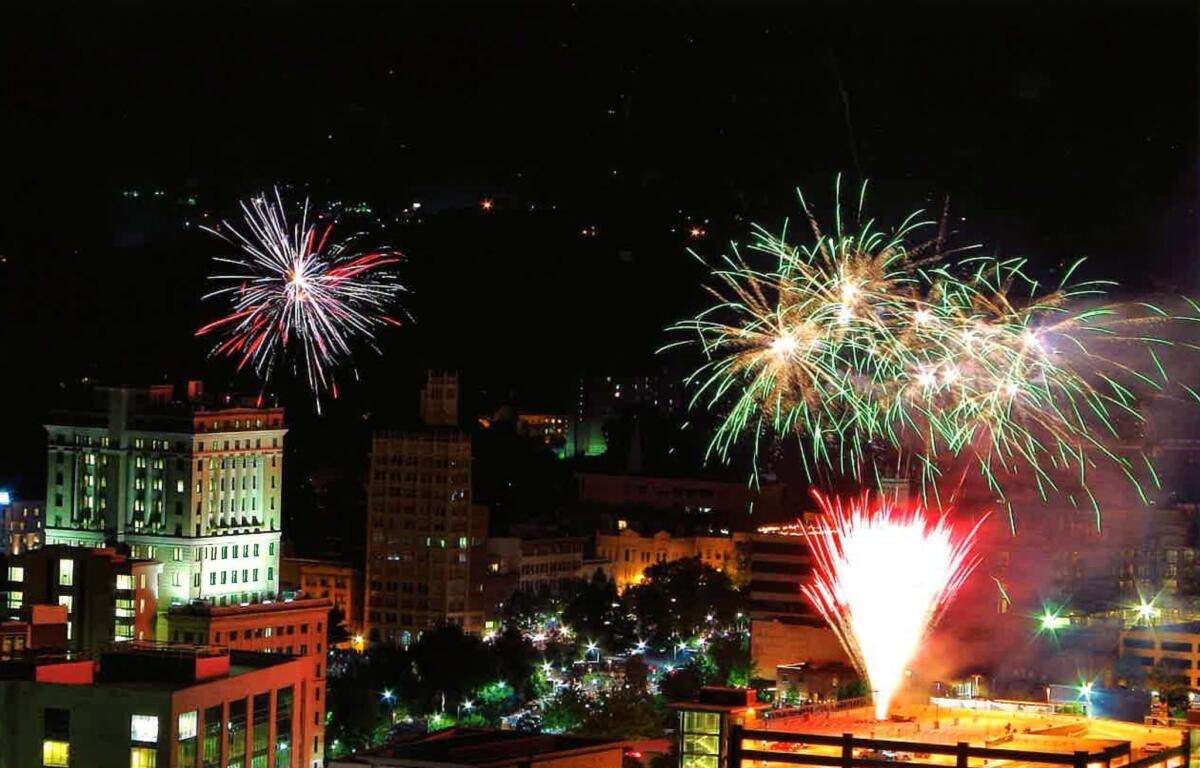 Night city skyline at night with colorful fireworks bursting above buildings; green and white bursts over the skyline and streetlights below.