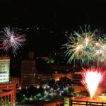 Night city skyline at night with colorful fireworks bursting above buildings; green and white bursts over the skyline and streetlights below.