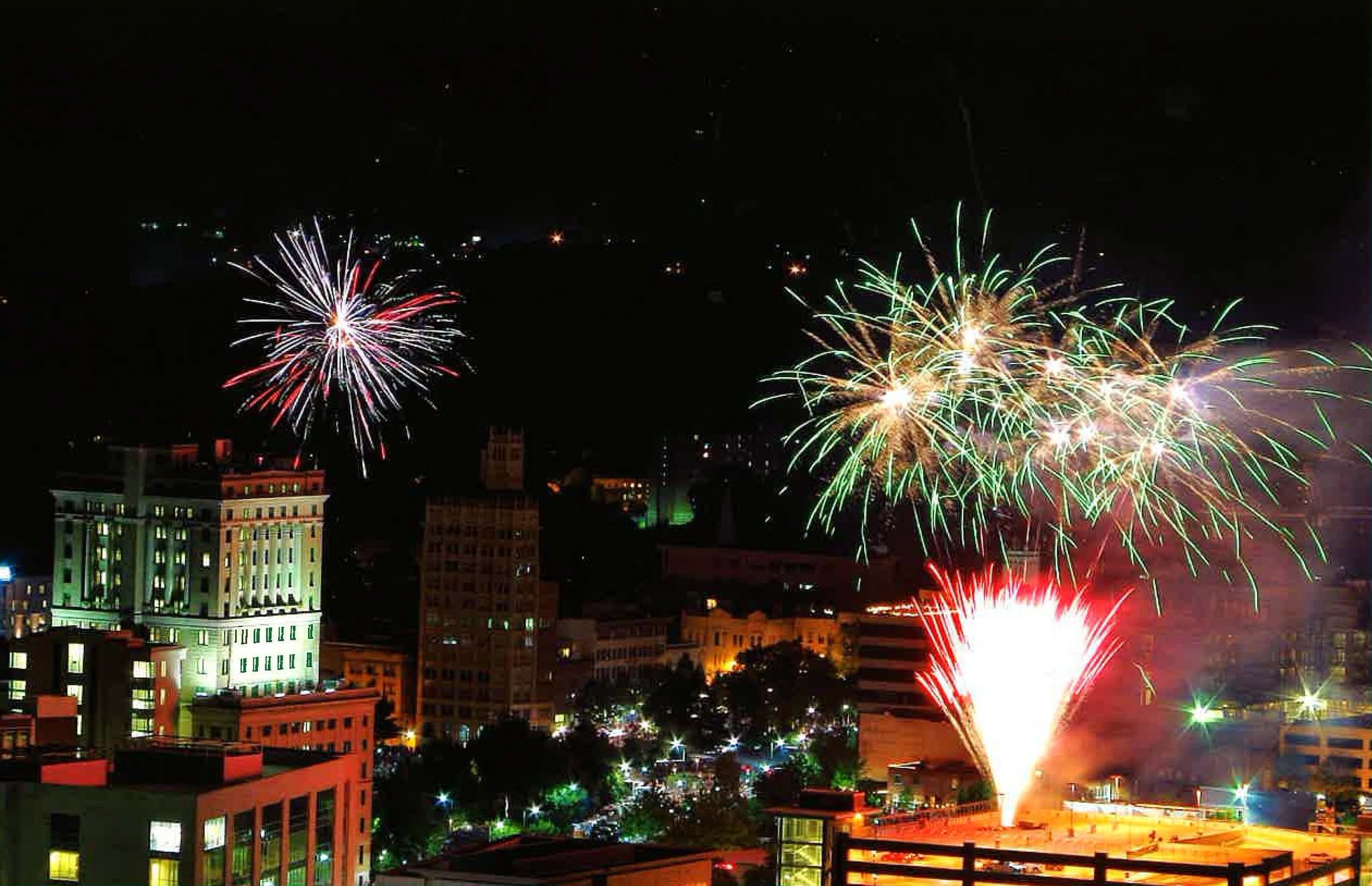 Night city skyline at night with colorful fireworks bursting above buildings; green and white bursts over the skyline and streetlights below.