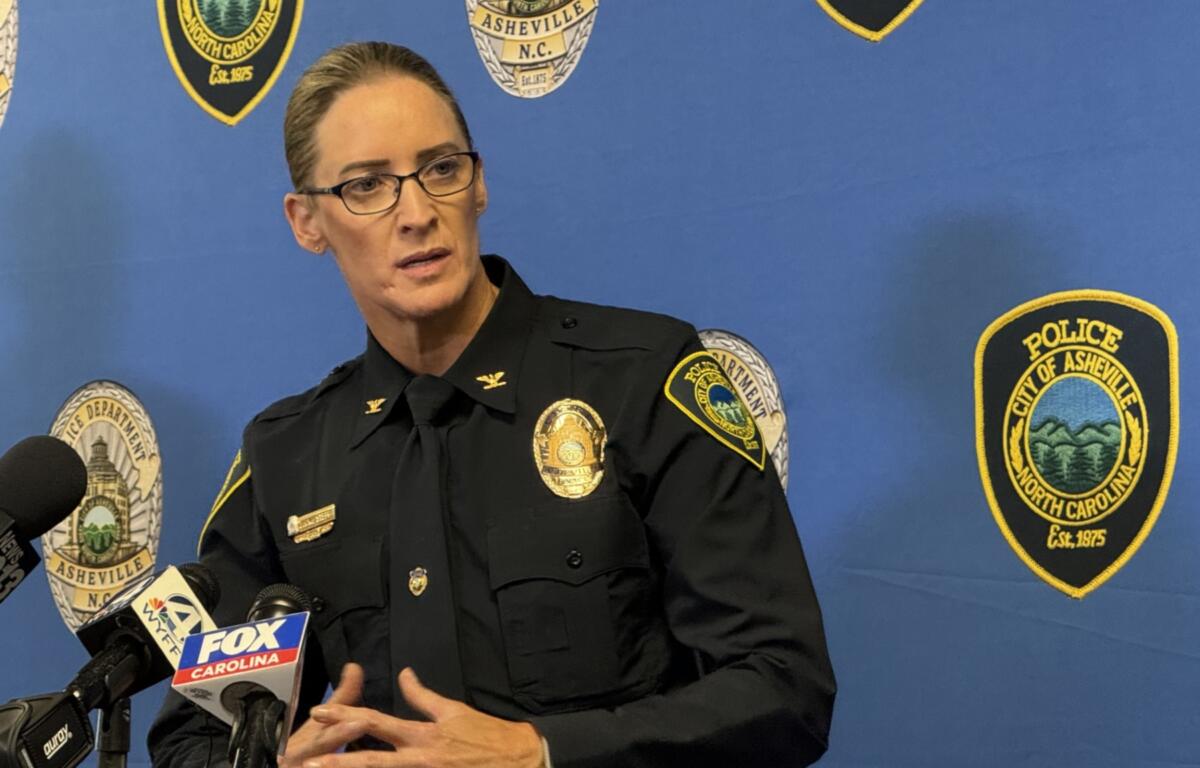 Female police officer in uniform speaks at a press conference, with microphones in front and a blue backdrop dotted with Asheville Police Department patches.