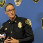 Female police officer in uniform speaks at a press conference, with microphones in front and a blue backdrop dotted with Asheville Police Department patches.